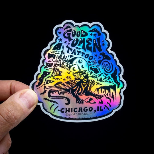 Holographic sticker with 'Good Omen Tattoo Chicago, IL' design held by a hand against a black background