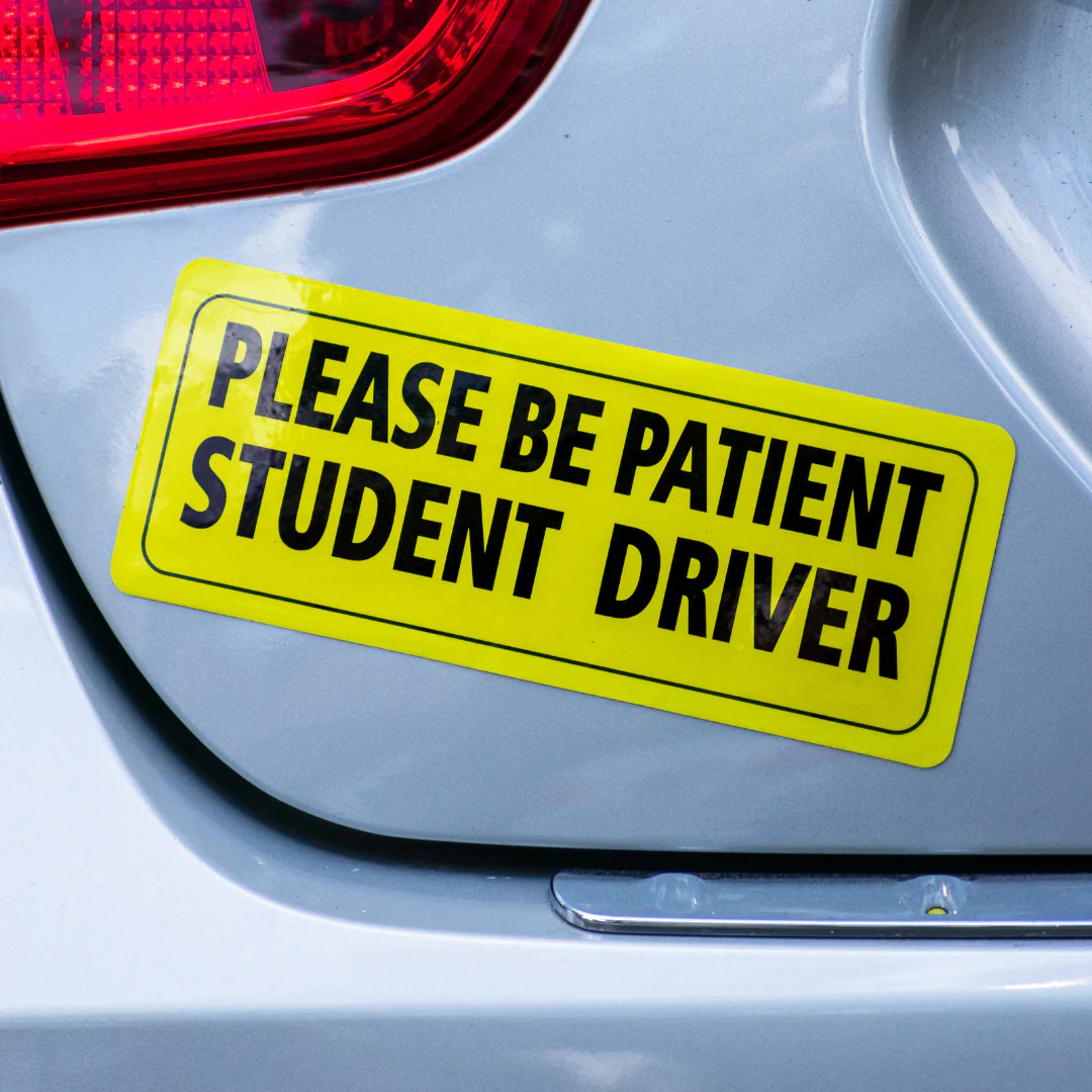 Yellow sign on a car tailgate reading 'PLEASE BE PATIENT STUDENT DRIVER' with a red light in the background.