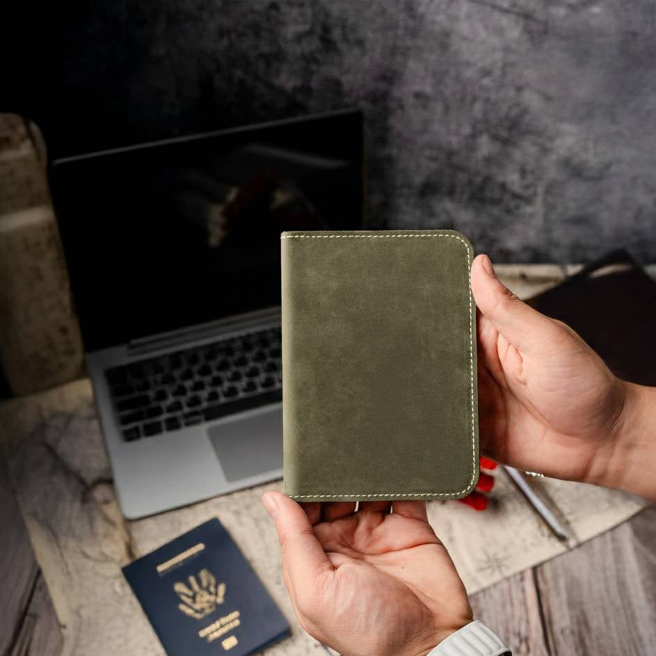 Hand holding a green leather passport holder with a laptop and passport on a wooden surface.