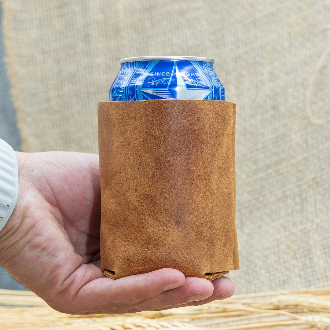 Hand holding a brown leather can cooler with a blue can inside against a neutral background