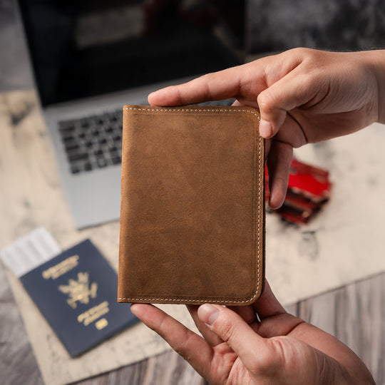 Brown leather wallet held by a hand with a blurred background of a laptop and passport.