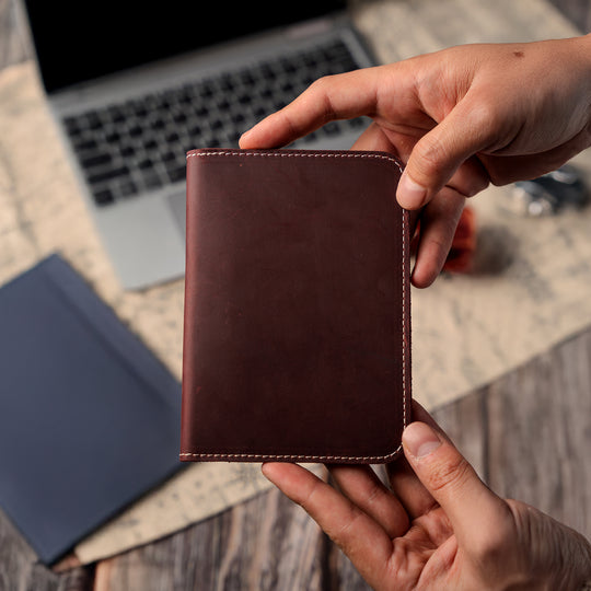 Person holding a brown leather wallet with a laptop and notebook in the background