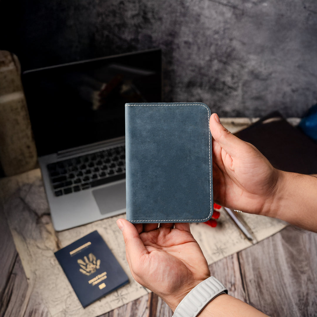 Hand holding a blue leather wallet with a laptop and passport on a wooden surface.